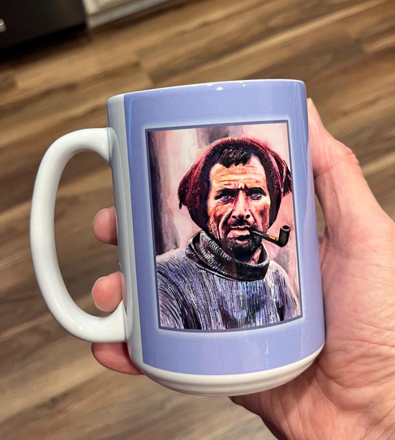 May include: A white ceramic mug with a blue background and a framed image of a man with a beard wearing a red hat and a pipe. The man is looking directly at the viewer.