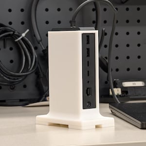 Peut inclure: Un support en plastique blanc avec un panneau noir comportant plusieurs ports. Le support a une base avec un bord arrondi et une forme rectangulaire.