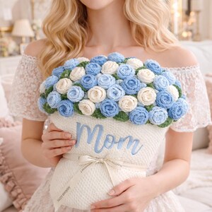 May include: A crocheted floral arrangement in a white container with the word "Mom" embroidered in blue. The arrangement features blue and cream-colored roses with green leaves. A white ribbon with text is tied around the container.