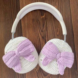 Puede incluir: Auriculares de crochet blanco con lazos morados en cada auricular. Los auriculares est&aacute;n sobre una superficie de madera marr&oacute;n.
