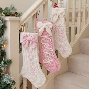 Könnte beinhalten: Drei Weihnachtsstrümpfe aus Zopfstrick mit Schleifenakzenten hängen an einer Treppe. Ein rosa Strumpf hat den Namen "Kelly" in Weiß gestickt. Die anderen beiden sind weiß mit rosa Stickerei. Links ist eine Weihnachtsbaumgirlande zu sehen.