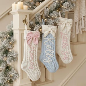 Puede incluir: Tres calcetines navideños de punto trenzado en blanco y azul claro, cada uno con un lazo en la parte superior. Los calcetines blancos tienen nombres bordados en rosa, mientras que el calcetín azul tiene un nombre bordado en blanco. Los calcetines cuelgan de una escalera decorada para las fiestas.