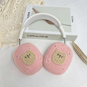 K&ouml;nnte beinhalten: Rosa geh&auml;kelte Ohrensch&uuml;tzer mit einem braunen B&auml;renkopf-Design. Die Ohrensch&uuml;tzer sind an einem wei&szlig;en Stirnband befestigt.