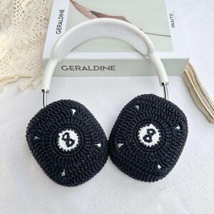 Puede incluir: Fundas para auriculares de crochet negro con un dise&ntilde;o de bola 8 blanca. Las fundas est&aacute;n unidas a una diadema blanca.