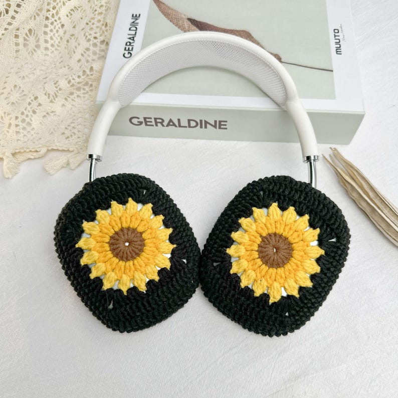 May include: White headphones with black crocheted earmuffs featuring sunflower designs. The earmuffs are made with yellow, brown, and black yarn.