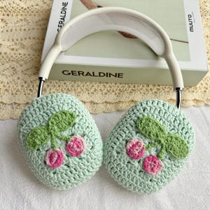 May include: Green crocheted headphone covers with a cherry design. The covers are attached to a white headband.