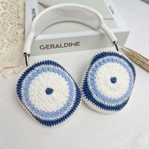Puede incluir: Fundas para auriculares de crochet blanco con rayas azules y azul claro. Las fundas tienen un patr&oacute;n circular en el centro.