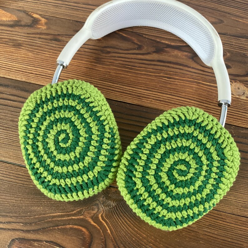 May include: Green and dark green crocheted earmuffs for headphones. The earmuffs are made with a circular pattern.