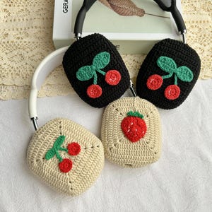 May include: Three crocheted headphone covers with different fruit designs. One cover features a red strawberry, while the other two feature red cherries with green leaves. The covers are made with a beige and black yarn.
