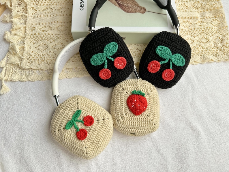 May include: Three crocheted headphone covers with different fruit designs. One cover features a red strawberry, while the other two feature red cherries with green leaves. The covers are made with a beige and black yarn.