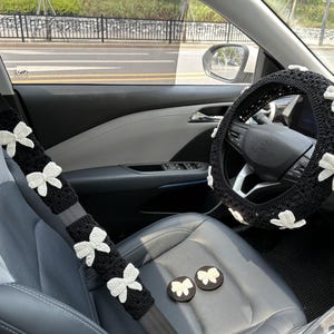 K&ouml;nnte beinhalten: Ein schwarzer Autoinnenraum mit wei&szlig;en geh&auml;kelten Schleifen auf den Sicherheitsgurtbez&uuml;gen und dem Lenkradbezug. Die Schleifen haben die Form von Schmetterlingen.