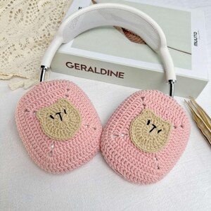 K&ouml;nnte beinhalten: Rosa geh&auml;kelte Ohrensch&uuml;tzer mit einem braunen B&auml;renkopf-Design. Die Ohrensch&uuml;tzer sind an einem wei&szlig;en Kopfband befestigt.