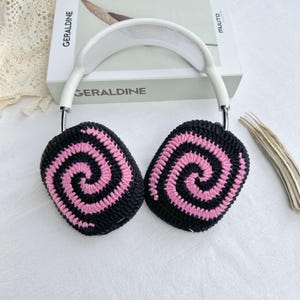 May include: White headphones with black and pink crocheted earmuffs featuring a spiral pattern. The earmuffs are attached to the headphones with a silver metal band.