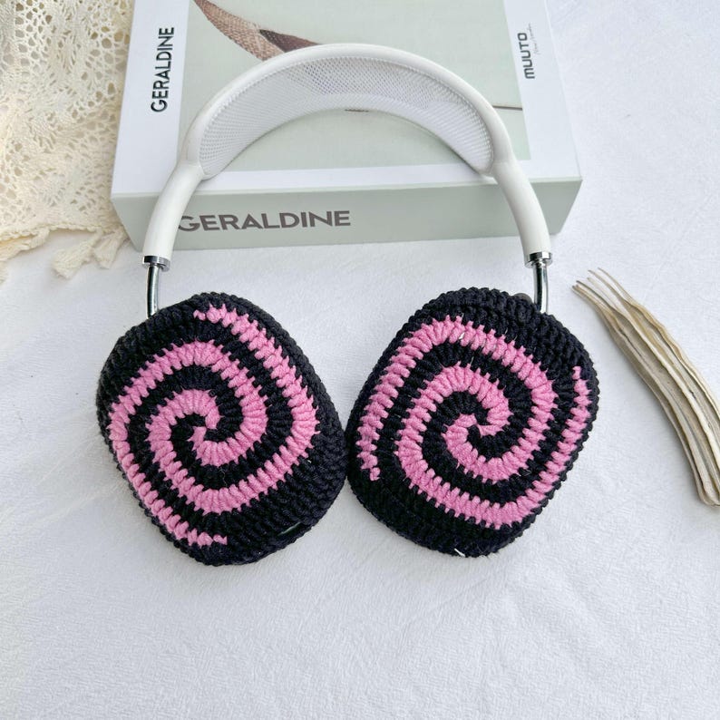 May include: White headphones with black and pink crocheted earmuffs featuring a spiral pattern. The earmuffs are attached to the headphones with a silver metal band.