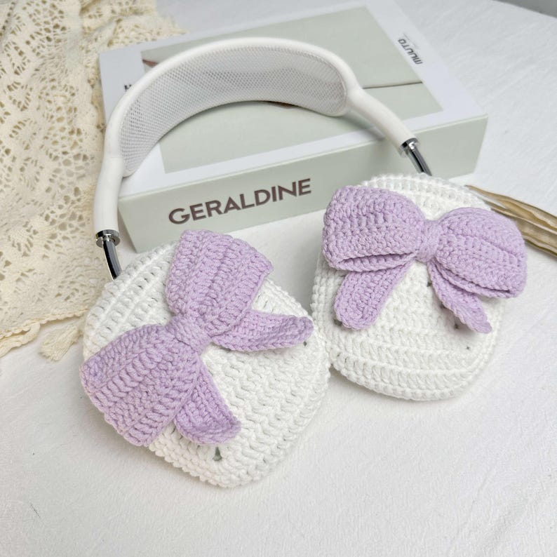 Puede incluir: Orejeras de ganchillo blancas con un lazo de ganchillo morado a cada lado. Las orejeras est&aacute;n unidas a una diadema blanca.