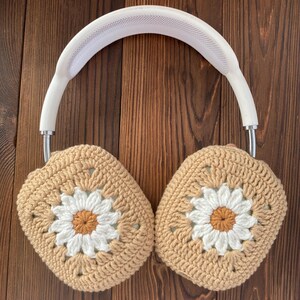 以下が含まれることがあります： 白いヘッドホンに、かぎ針編みで作った耳あてが付いています。耳あてはベージュで、白いデイジー模様が施されています。