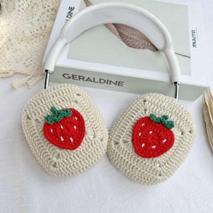 May include: White headphones with crocheted earmuffs featuring embroidered strawberry designs. The earmuffs are beige with red and green strawberries.