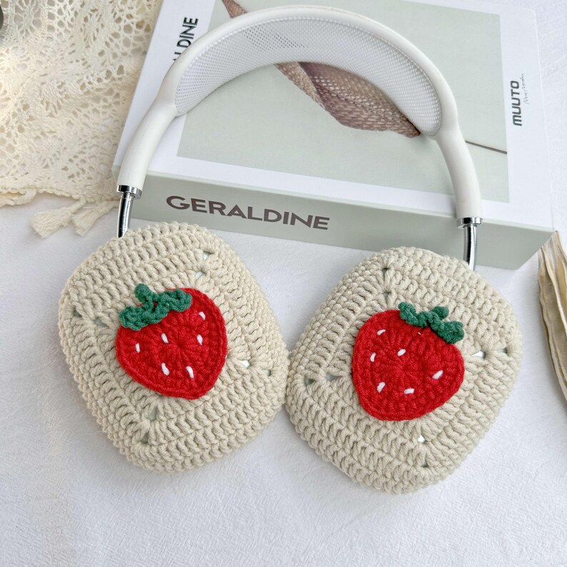 May include: White headphones with crocheted earmuffs featuring embroidered strawberry designs. The earmuffs are beige with red and green strawberries.