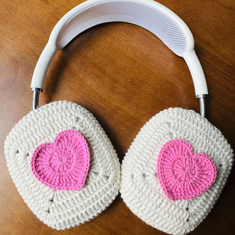 May include: White headphones with crocheted earmuffs featuring pink hearts. The earmuffs are made with a white yarn and have a pink heart crocheted on each earmuff.