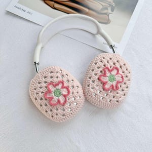 以下が含まれることがあります： 白いヘッドバンドと花柄のピンクのクロシェ編み耳あて。花はピンク、グリーン、ホワイトのアクセントになっています。