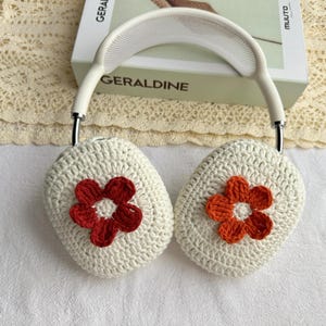 Puede incluir: Cubiertas de auriculares de crochet blanco con una flor roja y blanca en un lado y una flor naranja y blanca en el otro.