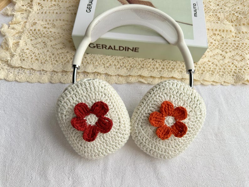 Puede incluir: Cubiertas de auriculares de crochet blanco con una flor roja y blanca en un lado y una flor naranja y blanca en el otro.