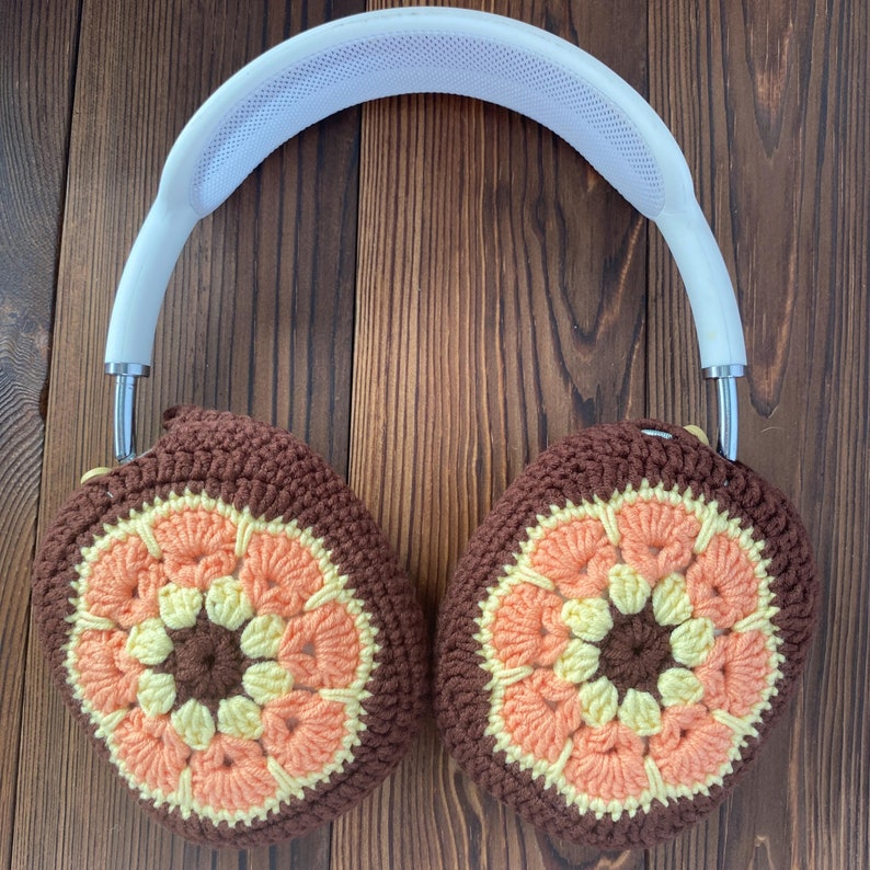 K&ouml;nnte beinhalten: Ein Paar geh&auml;kelte Kopfh&ouml;rerbez&uuml;ge mit einem braunen, orangefarbenen und gelben Blumenmuster. Die Bez&uuml;ge sind an einem wei&szlig;en Kopfband befestigt.