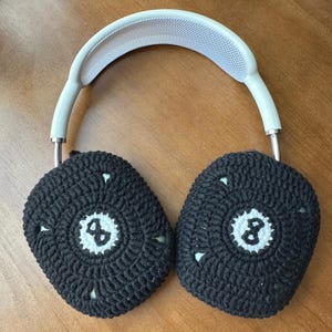 Puede incluir: Auriculares blancos de diadema con cubiertas de o&iacute;do de ganchillo negro. Cada cubierta de o&iacute;do tiene un c&iacute;rculo blanco con el n&uacute;mero "8" en el centro.