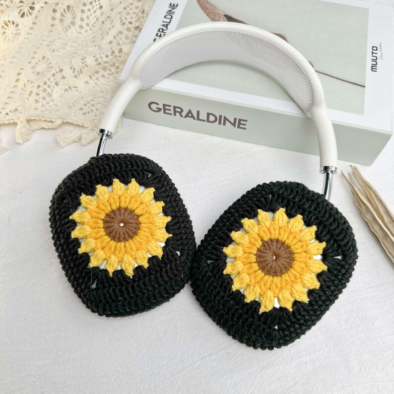 May include: White headphones with black crocheted earmuffs featuring yellow and brown sunflower designs. The earmuffs are crocheted with a square pattern.