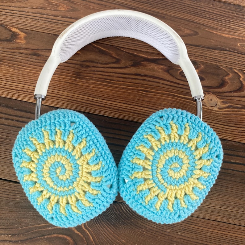 May include: White headphones with crocheted earmuffs. The earmuffs are blue and yellow with a sun-like pattern.