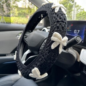 K&ouml;nnte beinhalten: Schwarze H&auml;kel-Lenkradabdeckung mit wei&szlig;en H&auml;kelb&ouml;gen. Die Abdeckung befindet sich auf einem Autolenkrad.