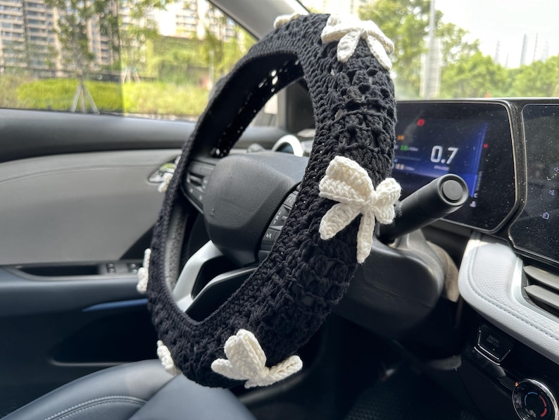 K&ouml;nnte beinhalten: Schwarze H&auml;kel-Lenkradabdeckung mit wei&szlig;en H&auml;kelb&ouml;gen. Die Abdeckung befindet sich auf einem Autolenkrad.