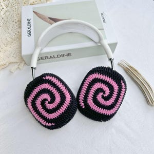 May include: White headband headphones with pink and black crocheted earmuffs featuring a spiral pattern.