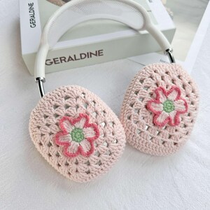 以下が含まれることがあります： 白いヘッドバンドとピンクの花柄のピンクのクロシェ耳あて。耳あては柔らかく、クロシェ編みの生地で作られており、繊細な花柄が施されています。