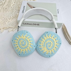 May include: White headphones with blue and yellow crocheted earmuffs featuring a sun design. The earmuffs are crocheted with a circular pattern.