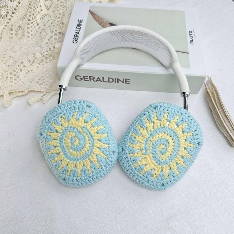 May include: White headphones with blue and yellow crocheted earmuffs featuring a sun design. The earmuffs are crocheted with a circular pattern.