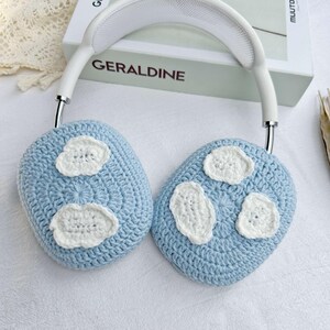 Puede incluir: Un par de orejeras de crochet azul claro con dise&ntilde;os de nubes de crochet blanco. Las orejeras est&aacute;n unidas a una diadema blanca con una banda de metal plateado.