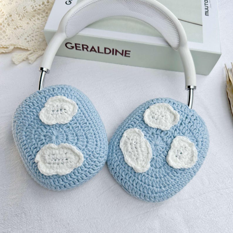 Puede incluir: Un par de orejeras de crochet azul claro con dise&ntilde;os de nubes de crochet blanco. Las orejeras est&aacute;n unidas a una diadema blanca con una banda de metal plateado.