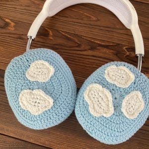 Puede incluir: Un par de orejeras de crochet azul claro con dise&ntilde;os en forma de nube blanca. Las orejeras est&aacute;n unidas a una diadema blanca.