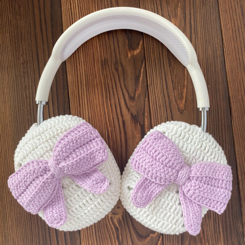 Puede incluir: Auriculares de crochet blanco con lazos morados en cada auricular. Los auriculares est&aacute;n sobre una superficie de madera marr&oacute;n.