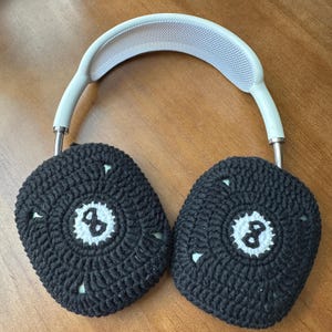 Puede incluir: Auriculares blancos con cubiertas de orejas de crochet negro. Las cubiertas de las orejas tienen un c&iacute;rculo blanco con el n&uacute;mero 8 en el centro.