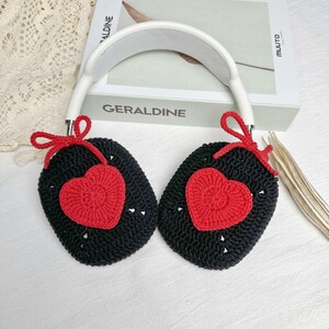 May include: Black crocheted earmuffs with red heart accents. The earmuffs are attached to a white headband.