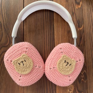 K&ouml;nnte beinhalten: Rosa gestrickte Kopfh&ouml;rerabdeckungen mit einem braunen geh&auml;kelten B&auml;renkopf auf jeder Seite. Die Kopfh&ouml;rerabdeckungen sind an einem wei&szlig;en Kopfband befestigt.