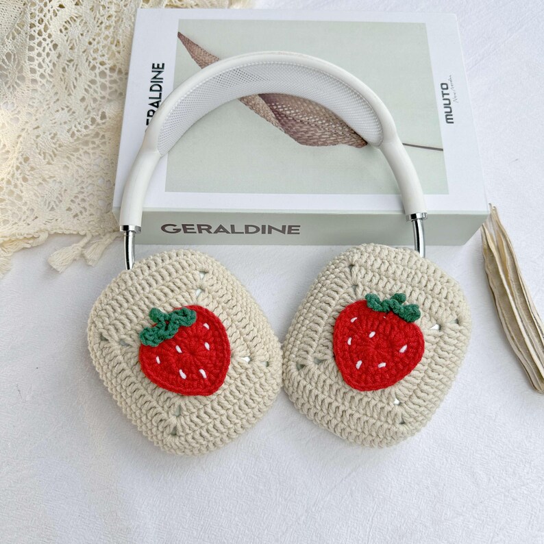 May include: White headphones with crocheted earmuffs featuring embroidered strawberry designs. The earmuffs are beige with red and green strawberries.