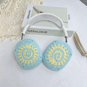 May include: White headband with blue and yellow crocheted earmuffs featuring a sun design. The earmuffs are made with a crocheted pattern of circles.