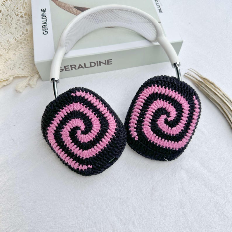 May include: White headphones with black and pink crocheted earmuffs featuring a spiral pattern. The earmuffs are handmade and add a unique touch to the headphones.