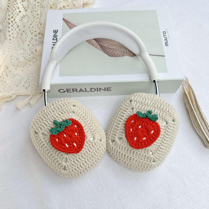 May include: White headphones with crocheted earmuffs featuring a strawberry design. The earmuffs are beige with red and green embroidered strawberries.