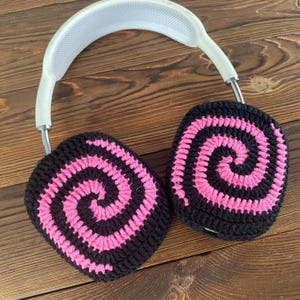 May include: Black and pink crocheted earmuffs with a spiral pattern. The earmuffs are attached to a white headband.
