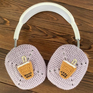 May include: Purple crocheted earmuffs with a white headband. Each earmuff has a brown and yellow embroidered boba tea cup with black boba pearls.