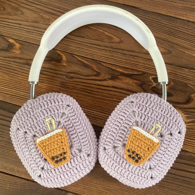 May include: Purple crocheted earmuffs with a white headband. Each earmuff has a brown and yellow embroidered boba tea cup with black boba pearls.